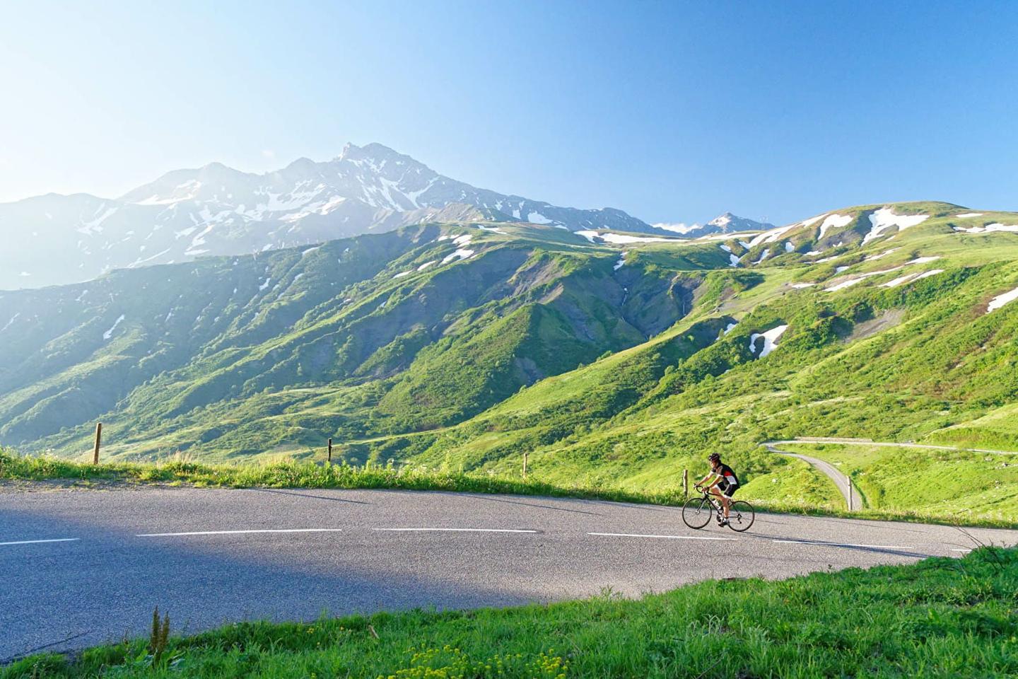 A cyclist climbing a winding road with lush green hills and snow-capped peaks in the background at Col de la Madeleine.