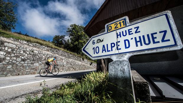 Solo cyclist near Alpe d'Huez road sign, iconic mountain pass in the French Alps, challenging climb scene.