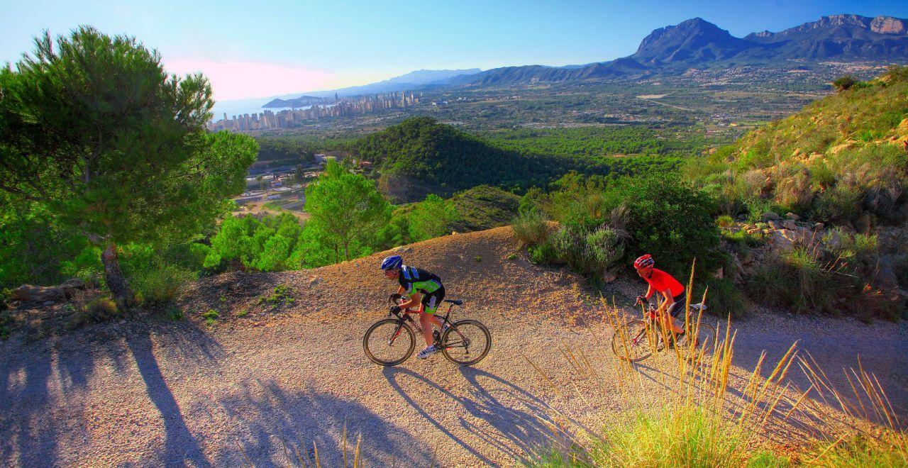 Two cyclists climbing a mountainous trail surrounded by lush greenery, with a distant view of Benidorm and the Mediterranean Sea