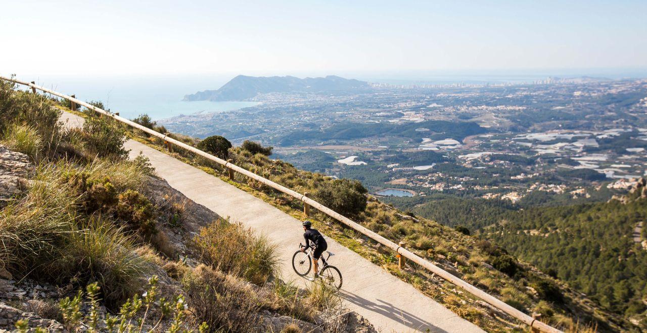 A lone cyclist ascending a winding mountain road with a panoramic view of the Mediterranean coastline and Benidorm city in the background