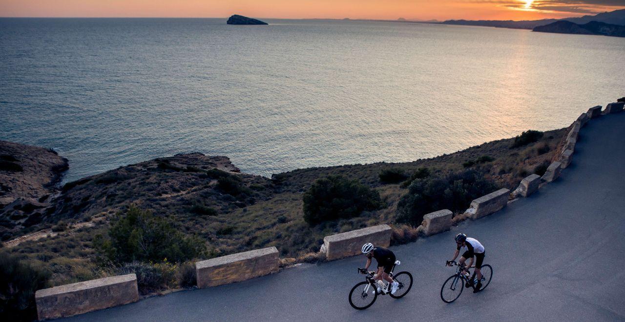 Two cyclists riding along a coastal road at sunset, with the sea and horizon bathed in golden light