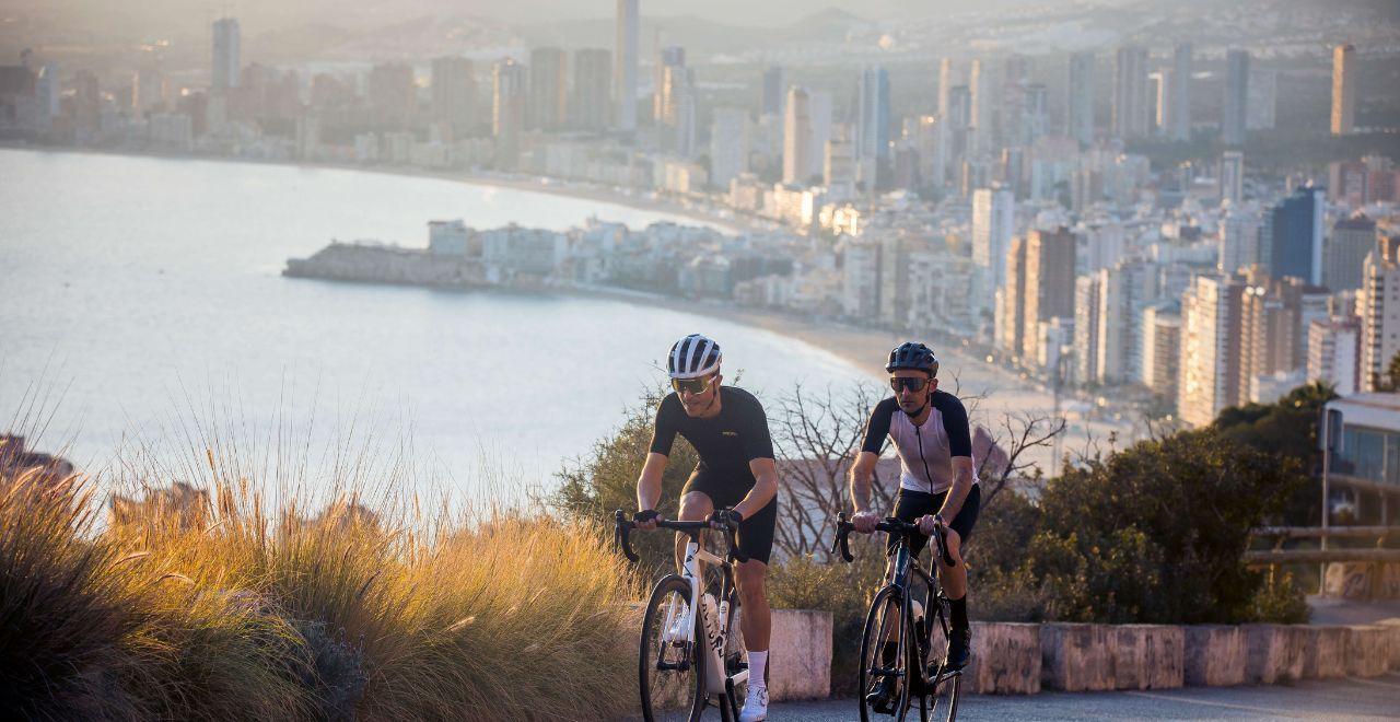 Two cyclists pedaling uphill with the high-rise buildings of Benidorm and the Mediterranean Sea in the background.