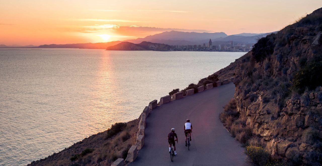 Two cyclists riding along a cliffside road at sunset, overlooking the calm sea and the silhouette of distant mountains