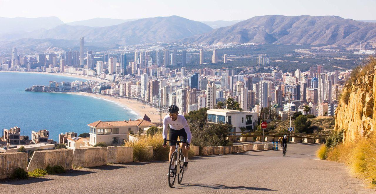 A cyclist pedaling uphill on a mountain road with a spectacular view of Benidorm's coastline and cityscape.