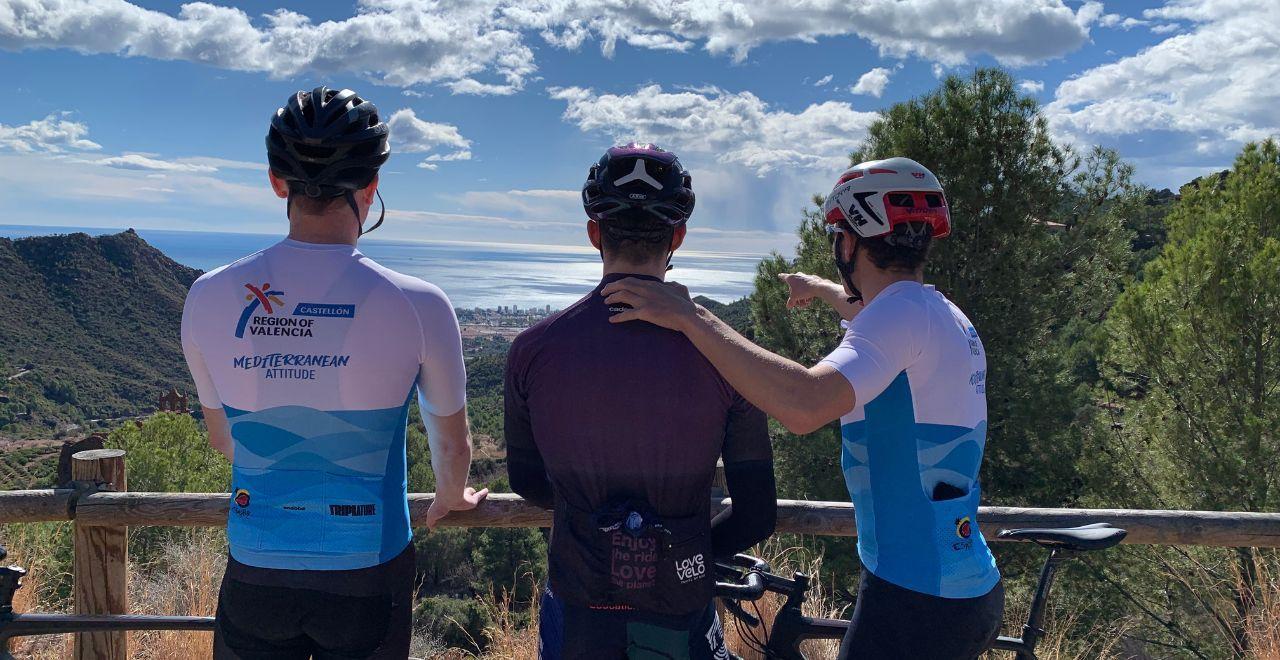 Three cyclists from the back, looking out over a mountainous landscape towards the sea and a city in the distance.