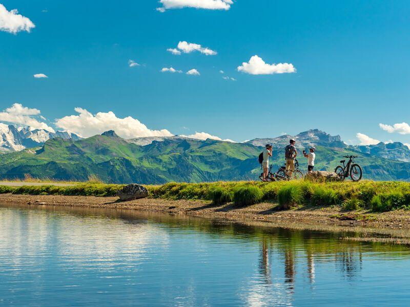 Cyclists standing by a lake with mountains in the background under a clear blue sky.