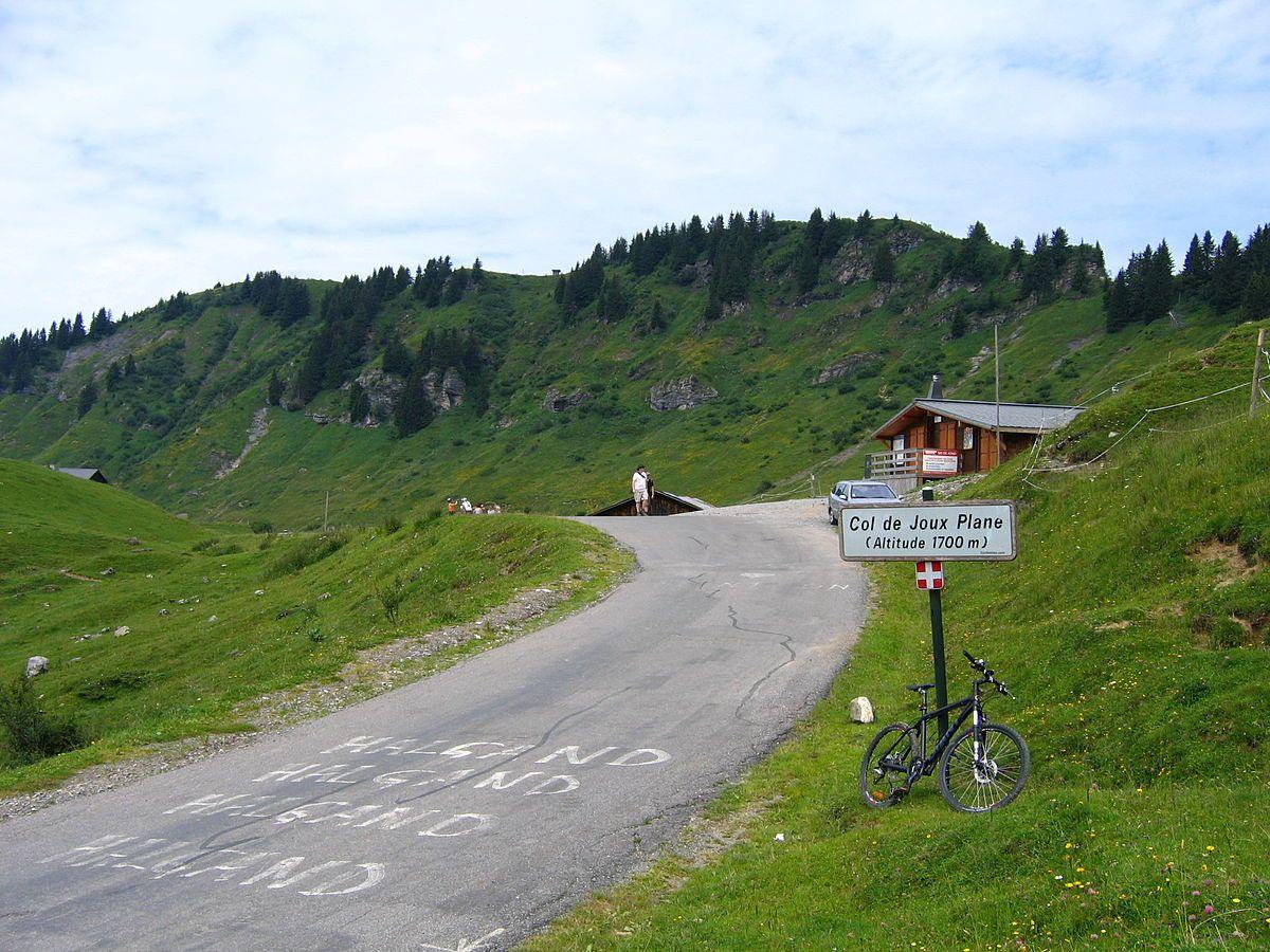 Bicycle leaning against a signpost at Col de Joux Plane with hills and a small building in the background.