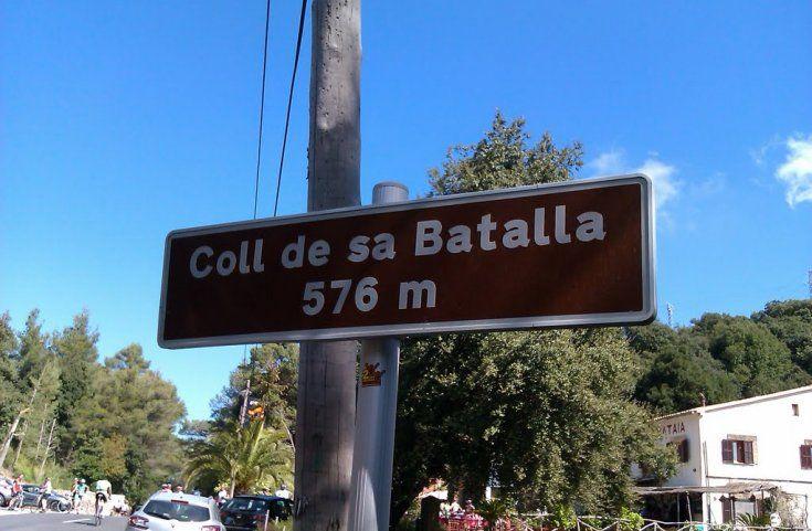Sign at the summit of Coll de sa Batalla in Mallorca, indicating an elevation of 576 meters, with cyclists and trees in the background.