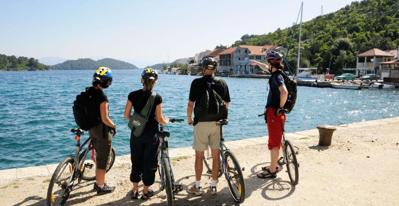 4 cyclists at the edge of a dock looking out on to a small harbour with mountains in the background