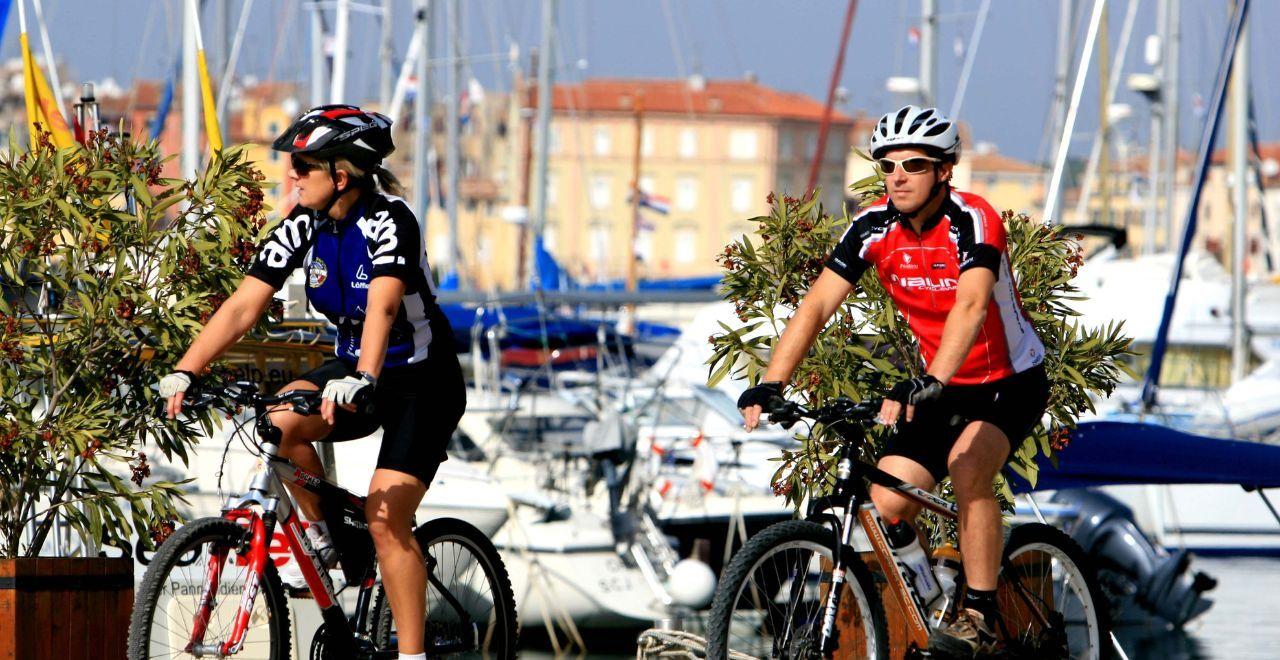 Cyclists riding beside a marina, enjoying a sunny day with views of sailboats, a perfect mix of cycling and nautical ambience