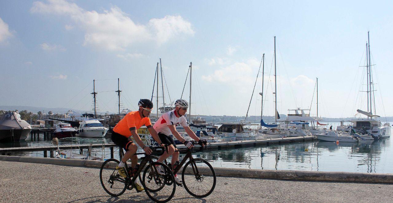 Cyclists enjoying a leisurely ride along a marina, with luxury yachts and clear blue skies, highlighting Cyprus's coastal charm for active travelers