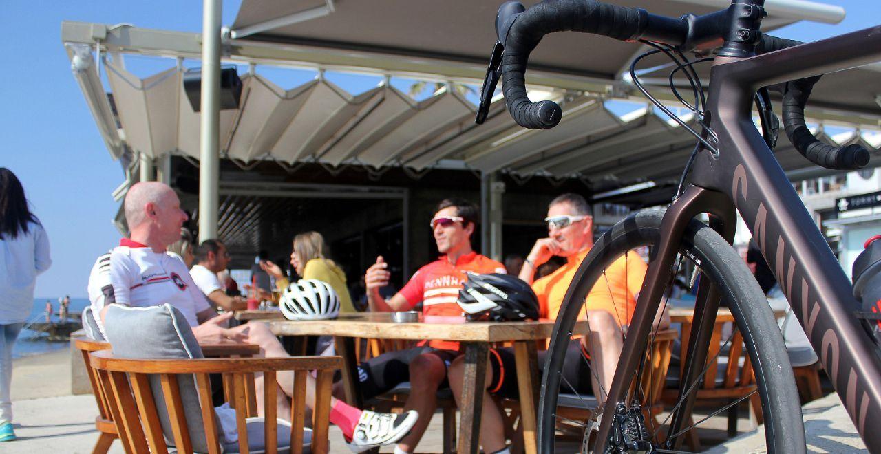 Cyclists enjoying a post-ride break at a seaside café in Cyprus, with bikes parked and picturesque views of the harbor, blending leisure with sport