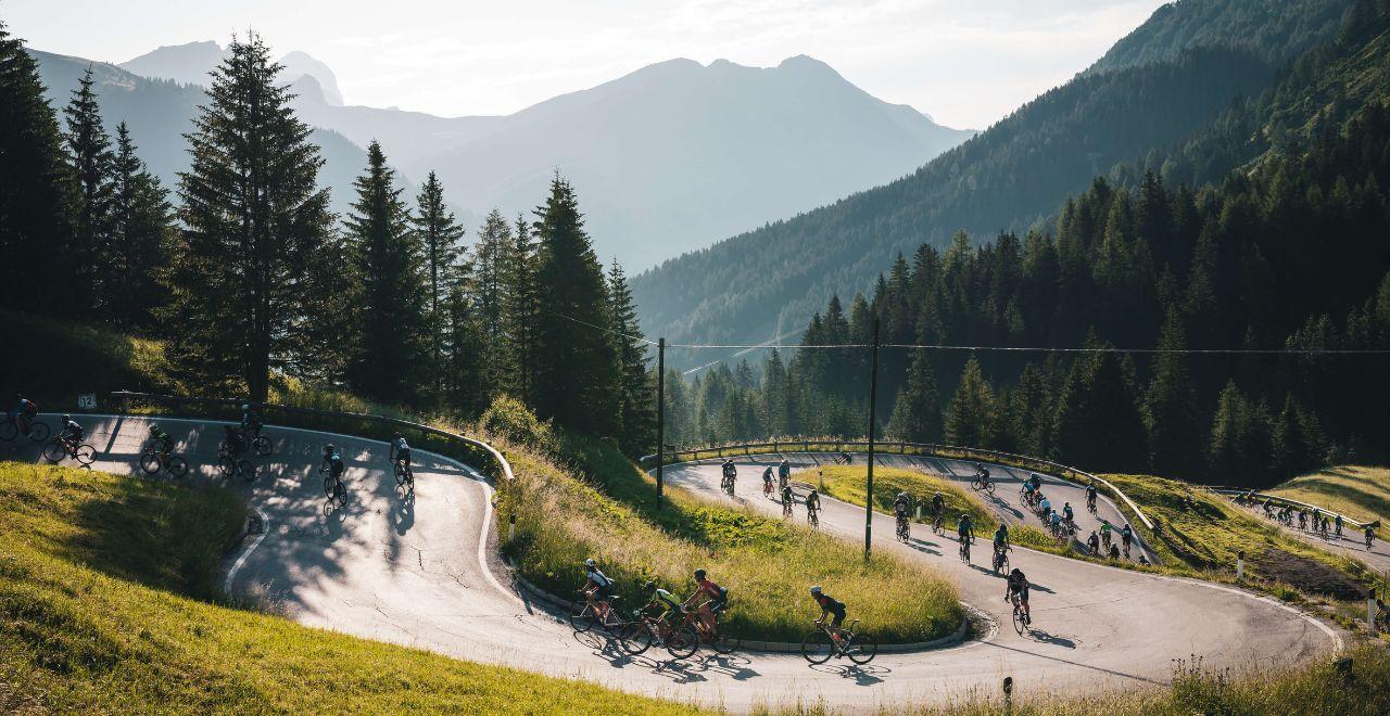 Cyclists navigate a hairpin turn on a mountain road in the Dolomites, the scene set against a backdrop of pine trees and distant peaks.