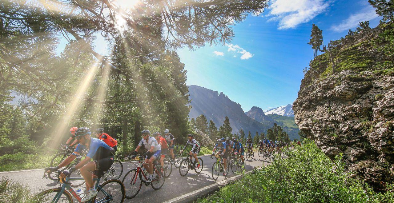 A group of cyclists ride through a forested area in the Dolomite mountains, sunlight filtering through the trees, with rocky peaks in the background.