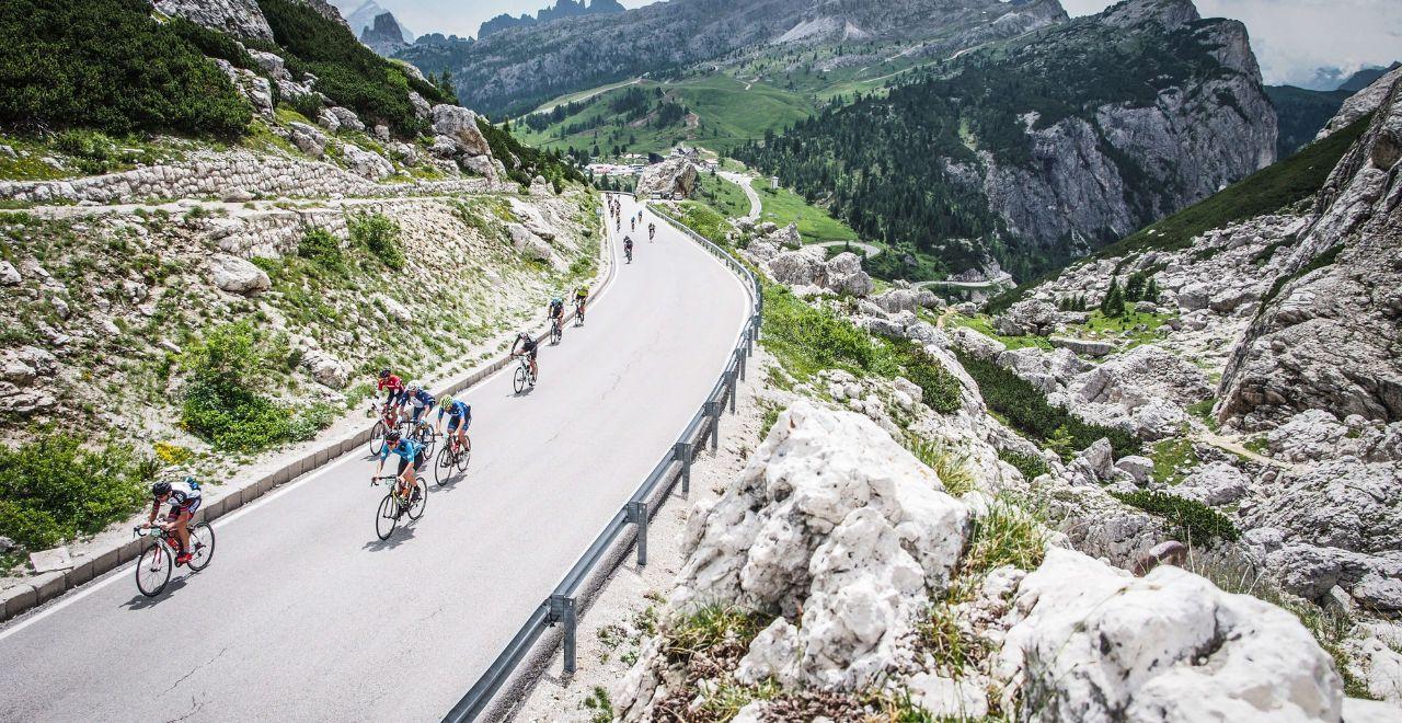 Cyclists race down a smooth, winding mountain road in the Dolomites, surrounded by rugged rocks and lush greenery.