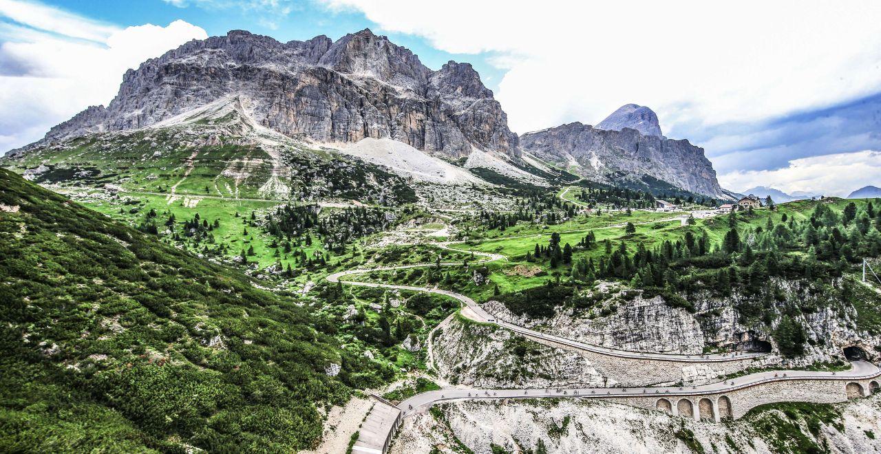 Cyclists navigate a winding road through the stunning Dolomite mountains, featuring rocky cliffs and verdant landscapes under a partly cloudy sky.
