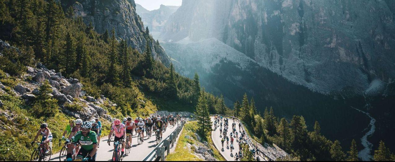 Cyclists participate in a road race through the scenic Dolomite mountains, surrounded by lush greenery and towering rock formations under a clear sky.