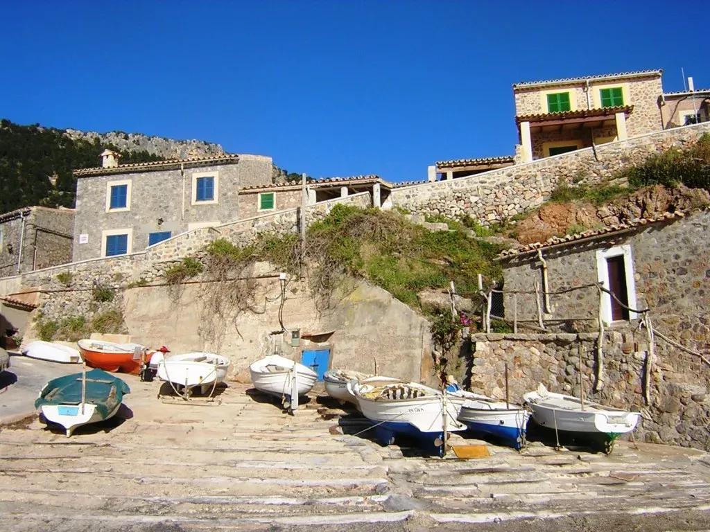 Small boats docked on a ramp leading to stone houses with green shutters in the quaint coastal village of Es Port de Valldemossa under a bright blue sky.