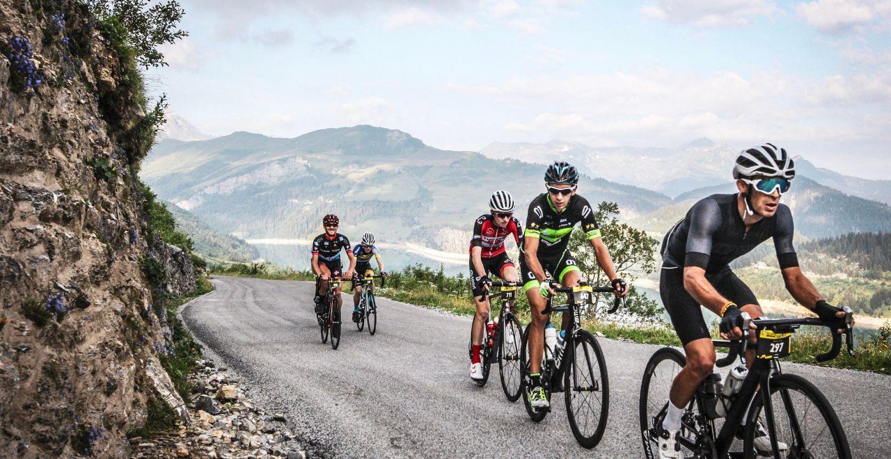 Group of cyclists riding on a winding mountain road with scenic views of hills and a lake.