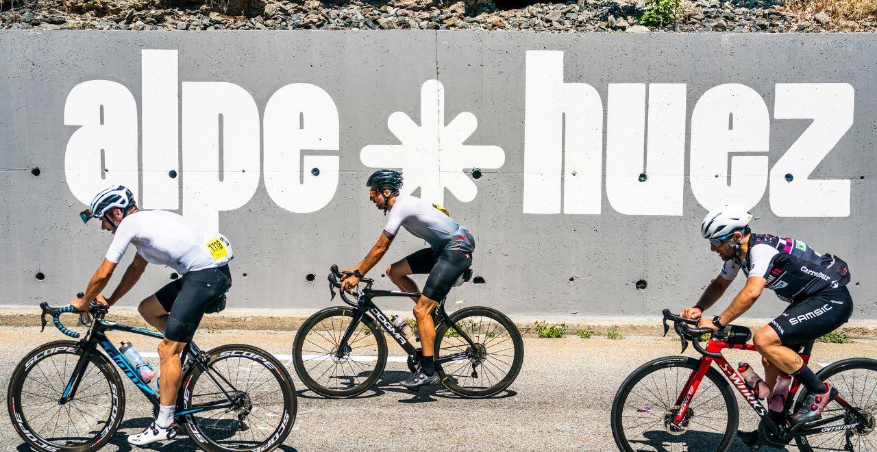 Cyclists riding past an Alpe d'Huez sign on a sunny day.