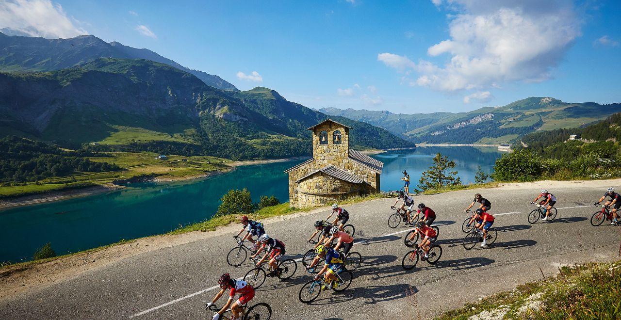 Cyclists passing a lakeside church on a mountain road.