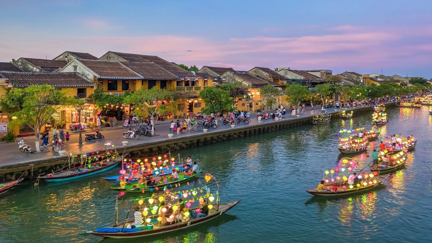 A scenic view of Hoi An ancient town at dusk, with vibrant lantern-lit boats floating on the river, historic buildings along the waterfront, and a lively atmosphere with people walking and riding motorcycles.