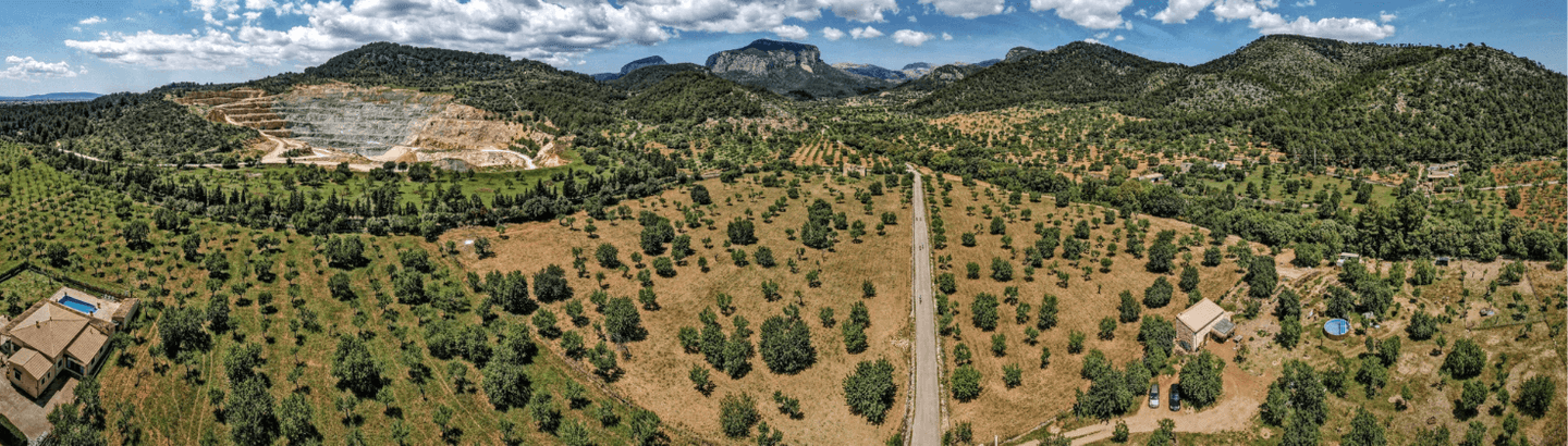 Aerial view of a rural landscape near Port Pollensa, Mallorca, featuring a road flanked by orchards and rocky hills with distant mountains.