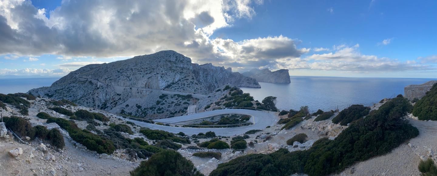 Panoramic view of the winding road to Cap Formentor, surrounded by rugged rocky cliffs and lush greenery, with the blue sea stretching out under a partly cloudy sky.