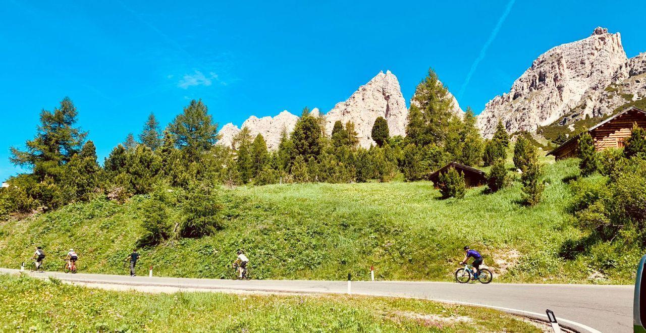 Cyclists riding on a mountain road with rocky peaks and pine trees in the Dolomites.