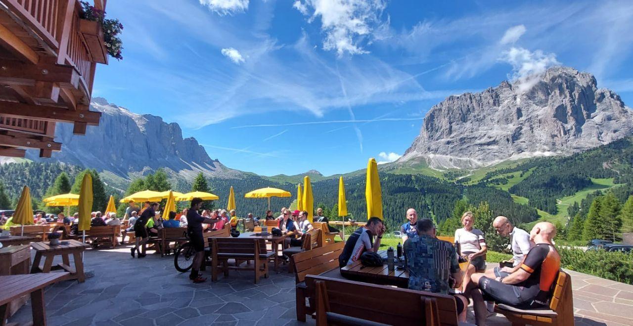 Outdoor dining with people under yellow umbrellas, Dolomites mountains in background.