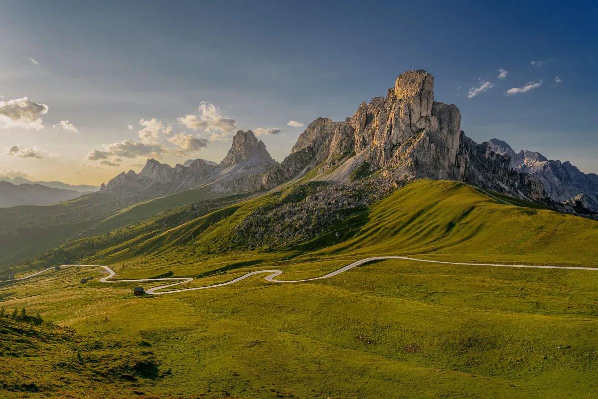 A winding road through Passo Giau in the Dolomites, with green alpine meadows leading up to dramatic rocky peaks under a clear blue sky.