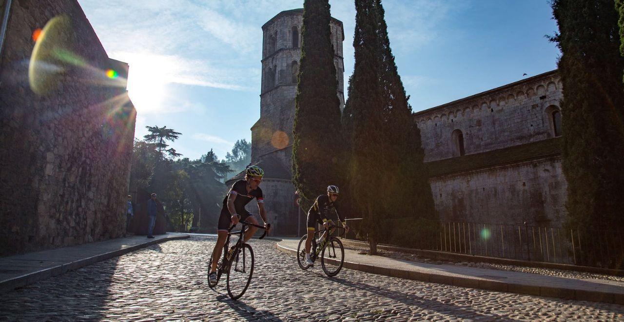 2 cyclists riding on cobbles through a medieval town under blue sky