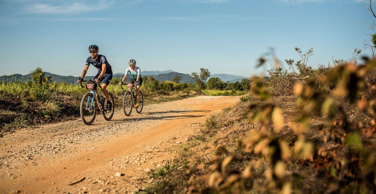Cyclists riding through a rural dirt path with fields and mountains.