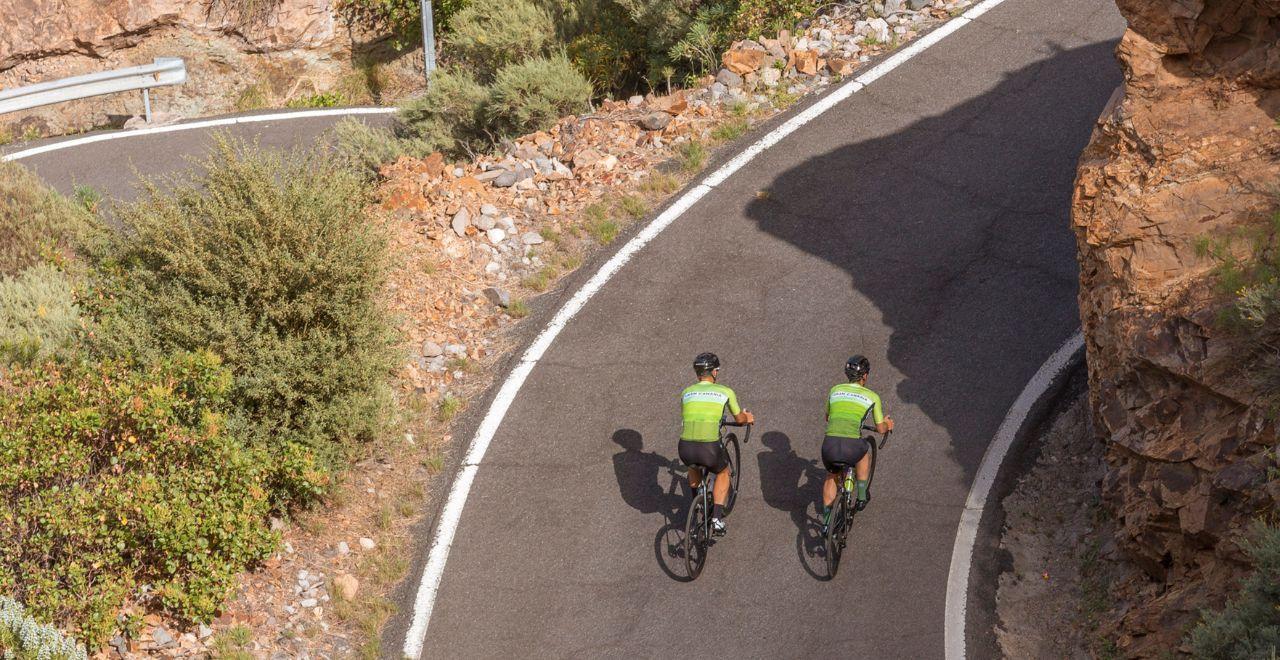 Two cyclists riding on a winding road with rocky cliffs.