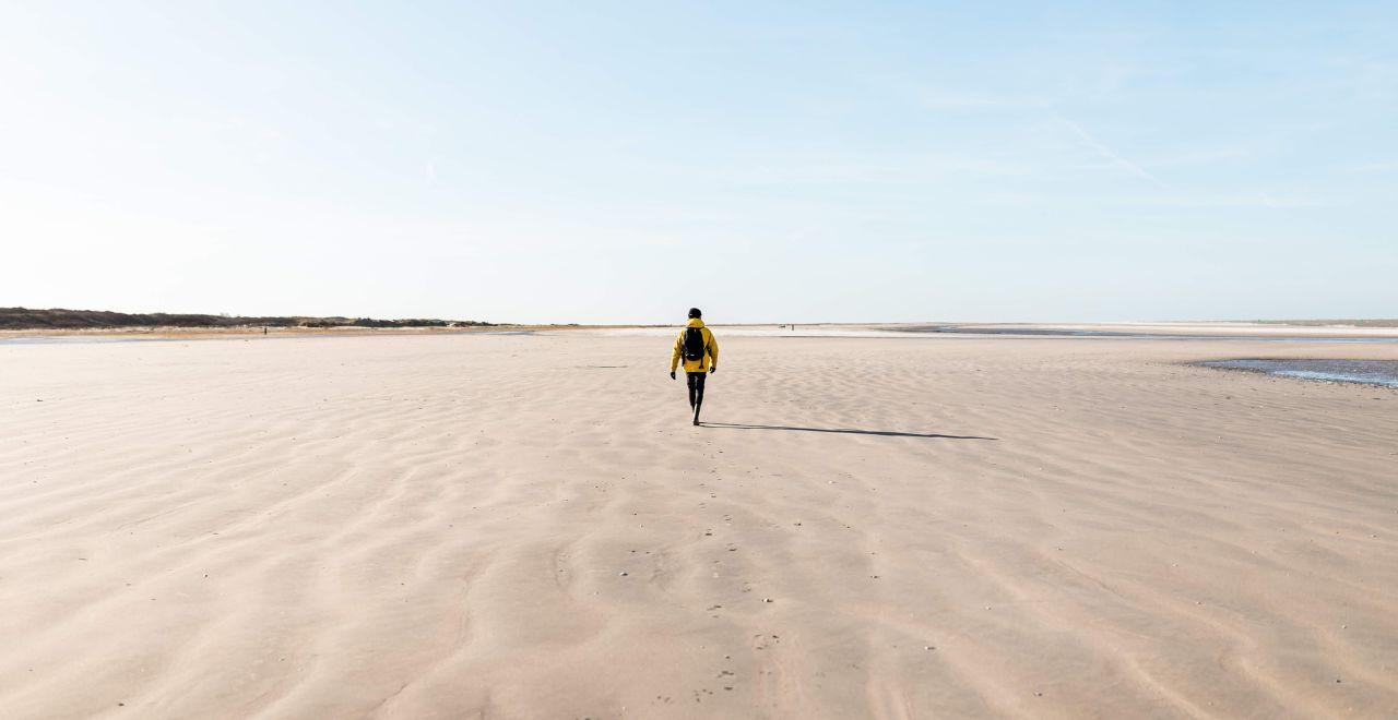 Person in a yellow jacket walking alone on a wide, empty beach with rippled sand
