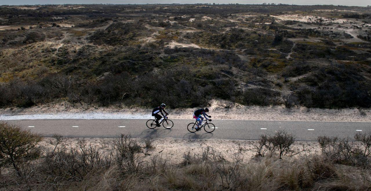 Two cyclists on a paved path through rugged sand dunes in a coastal landscape