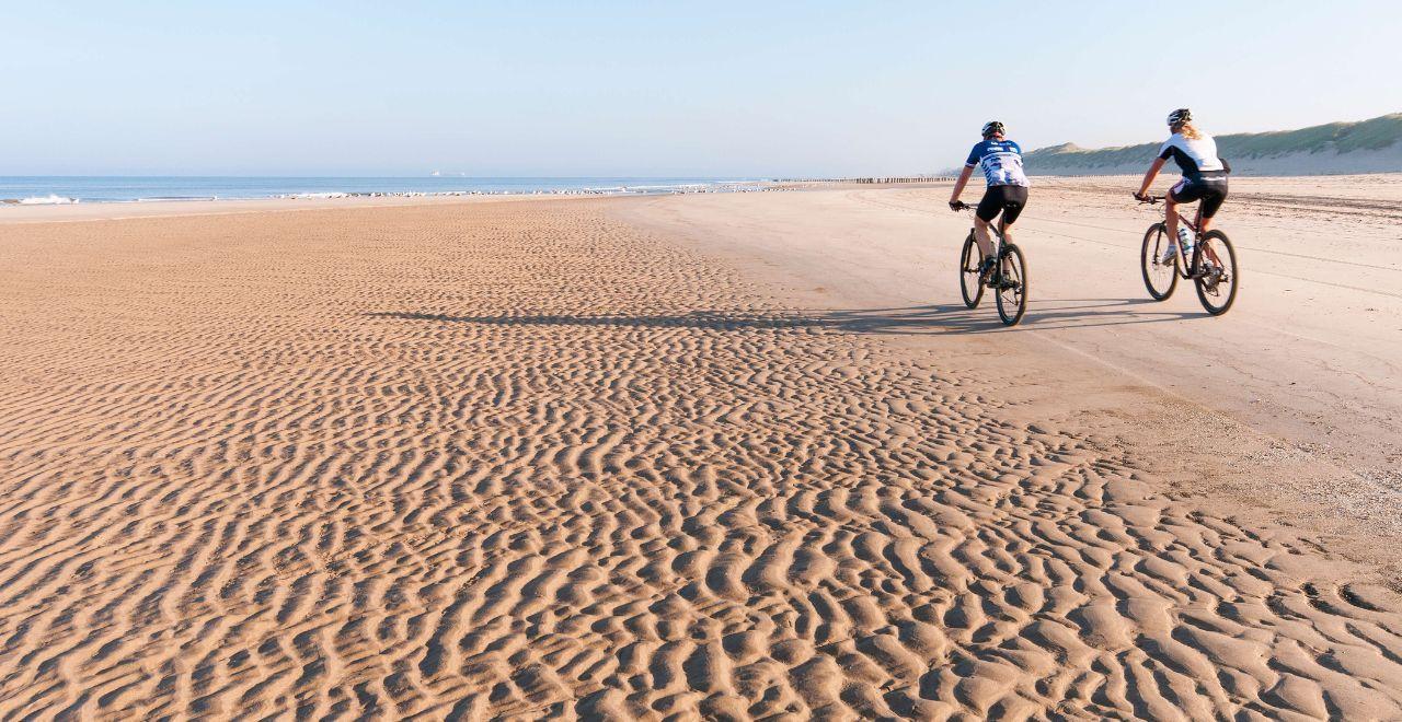 Two cyclists riding on a wide, sandy beach with gentle waves and dunes in the background