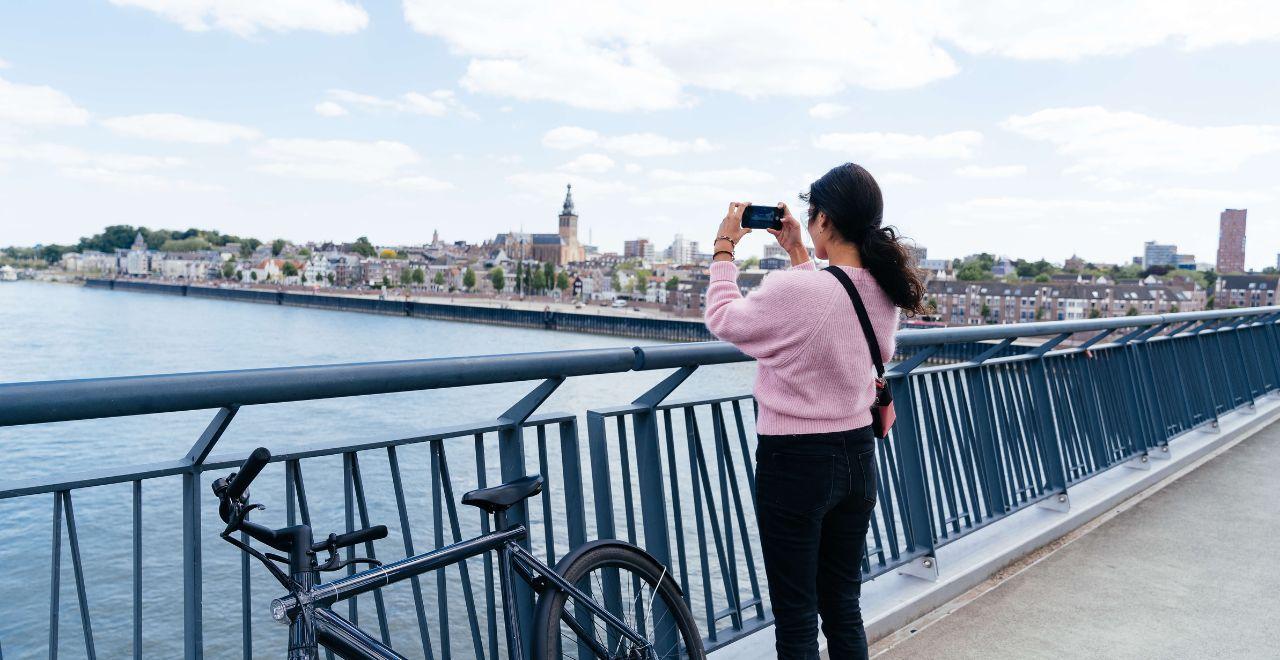 Woman taking a photo with her phone from a bridge overlooking a river and European cityscape