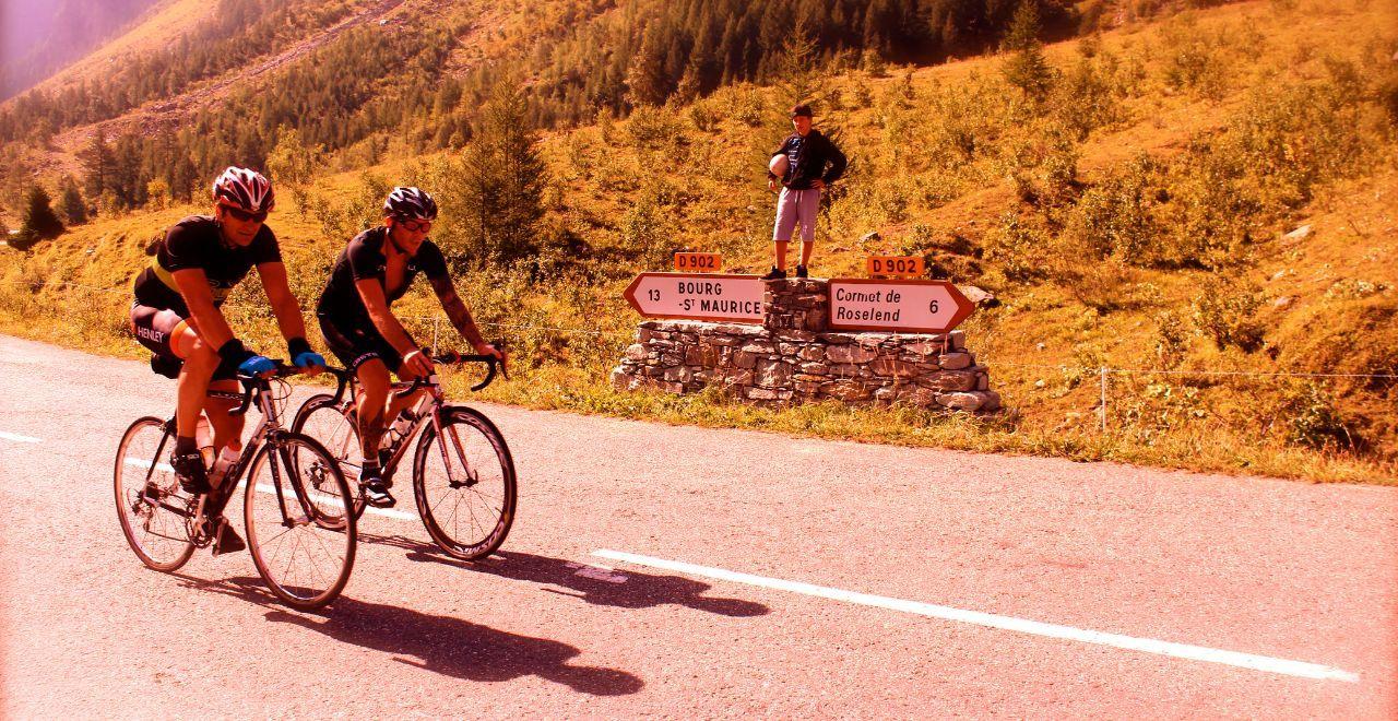 Two cyclists riding past a road sign showing directions to Bourg-Saint-Maurice and Cormet de Roselend.