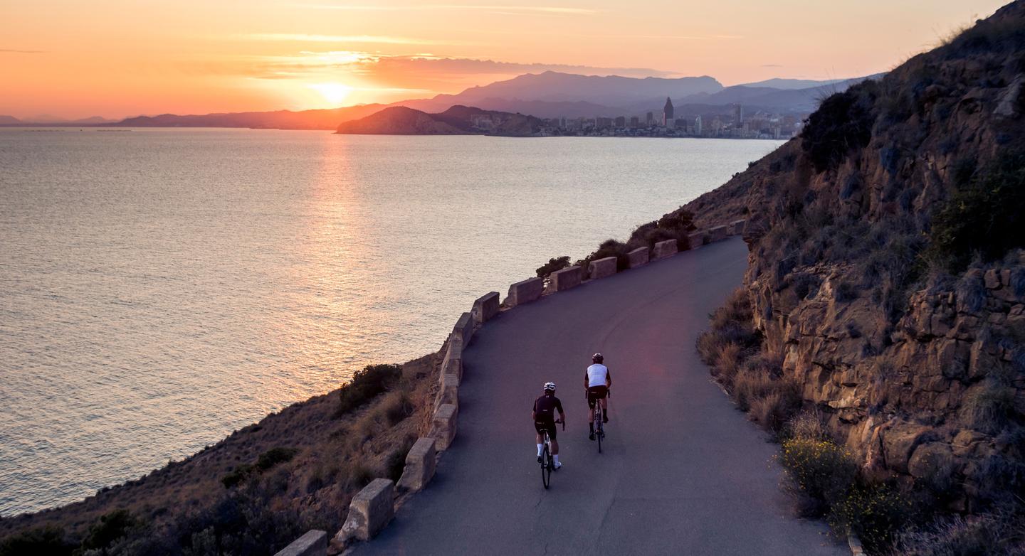 “Two cyclists riding uphill on a coastal road with a scenic view of the ocean and an island at sunset in Calpe, Costa Blanca.”
