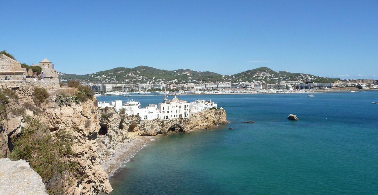 Coastal view of Ibiza with white buildings on rocky cliffs overlooking clear blue waters, with hills and a cityscape in the background.