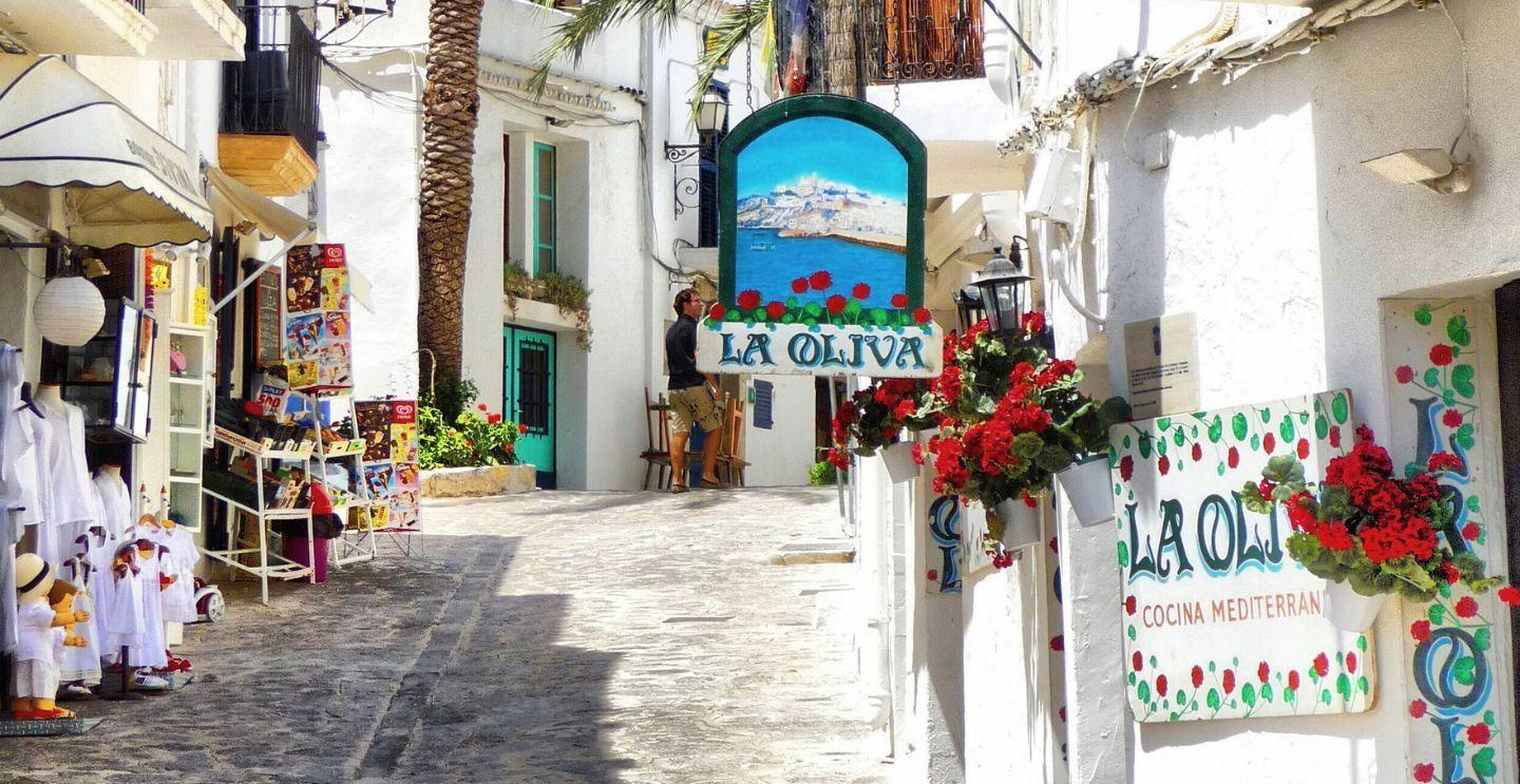  Quaint narrow street in Ibiza with whitewashed buildings, colorful storefronts, and a sign for La Oliva restaurant adorned with red flowers.
