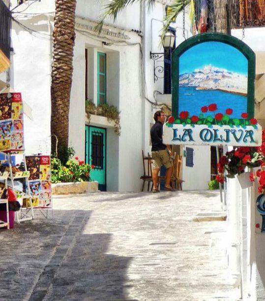 Charming white-washed alley with colorful pottery and a sign for 'La Oliva' restaurant in a sunny Mediterranean village