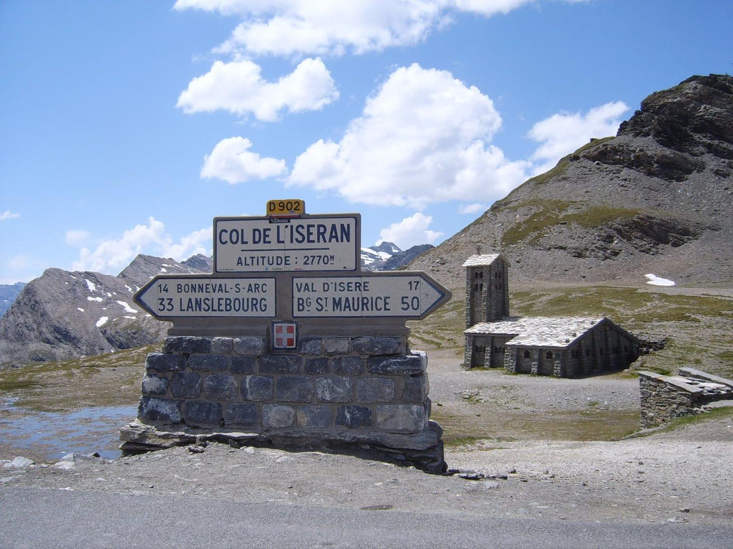 The summit sign of Col de l'Iseran at an altitude of 2770m, with a small stone chapel in the background and the surrounding Alpine landscape under a clear blue sky.