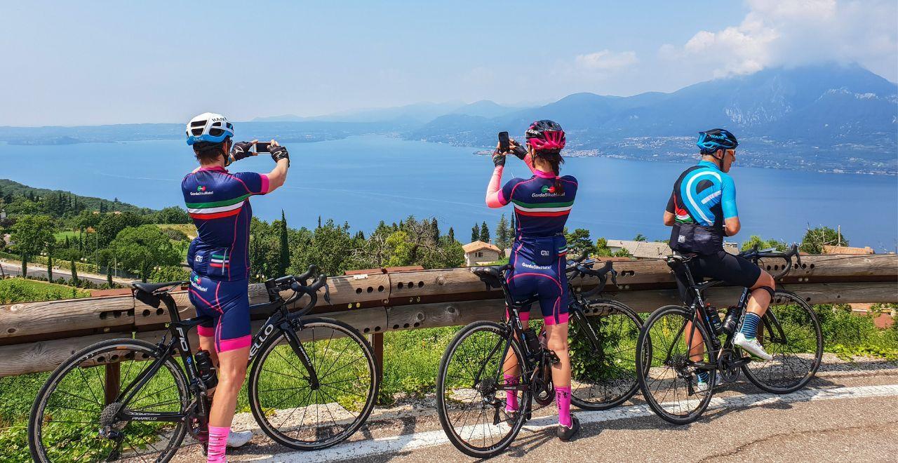 Three cyclists taking photos with their phones, overlooking a scenic lake and mountains