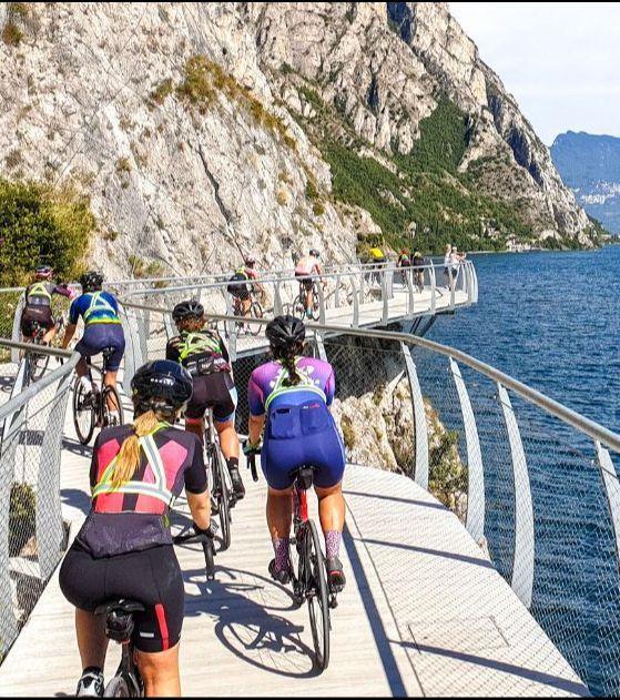 A group of cyclists on a narrow cycling path along the side of a rocky cliff overlooking a clear blue lake.
