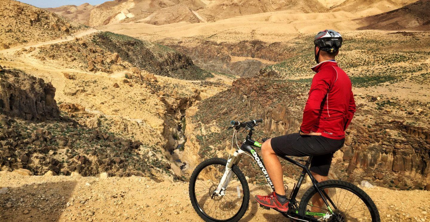Cyclist in a red jacket pausing to admire a rugged canyon landscape with hills and rocky 