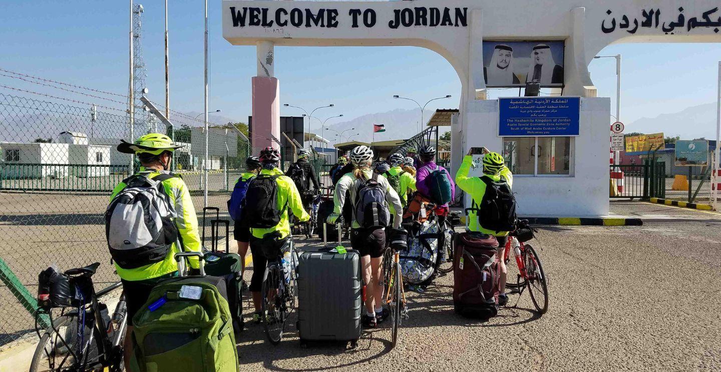 Group of cyclists with luggage gathered at the "Welcome to Jordan" border crossing sign.