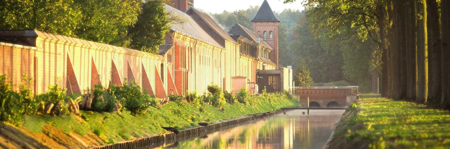 A serene canal in Belgium's Kempen region, bordered by historic brick buildings and lush greenery, bathed in golden sunlight. The tranquil water reflects the surrounding scenery, creating a picturesque and peaceful atmosphere.