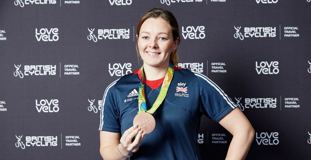 Female British cyclist holding a bronze medal in front of Love Velo backdrop